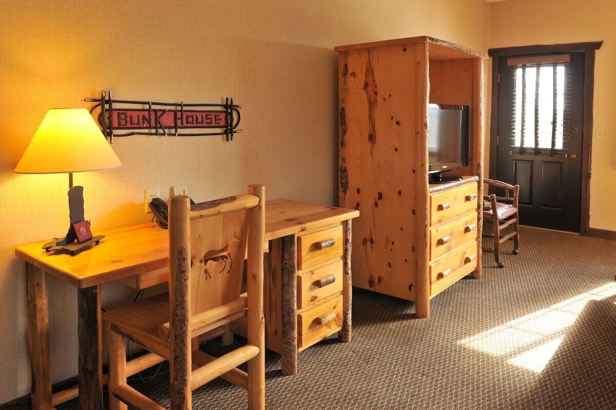 A rustic corner of the room features a wooden desk accompanied by a chair and a dresser with multiple drawers. A TV is housed in a unit made of natural wood. A slatted door allows natural light to enter the space, creating a welcoming atmosphere.