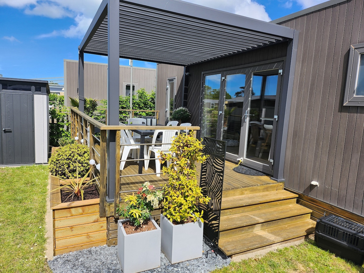 An outdoor wooden terrace is featured, equipped with a pergola providing shade. Garden furniture, including white chairs and a small table, is arranged for relaxation. Surrounding planters showcase greenery and flowering plants, creating a pleasant atmosphere. Steps lead from the terrace to the grassy area.