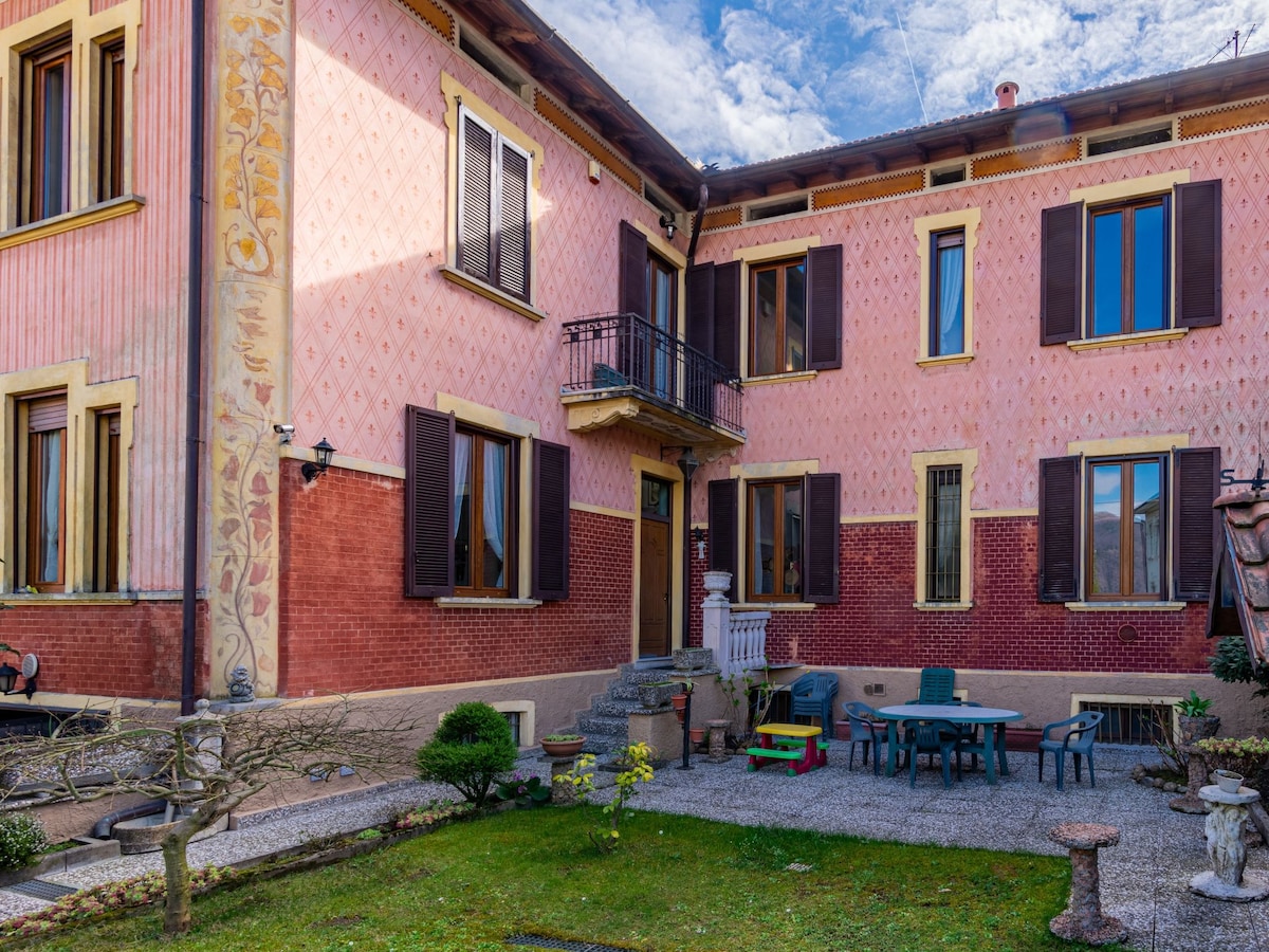 The exterior of a terraced house features a pink facade adorned with decorative elements. The entrance is framed by a small balcony, and a garden with gravel pathways is visible. Garden furniture is arranged around a small grassy area, complemented by ornamental plants.