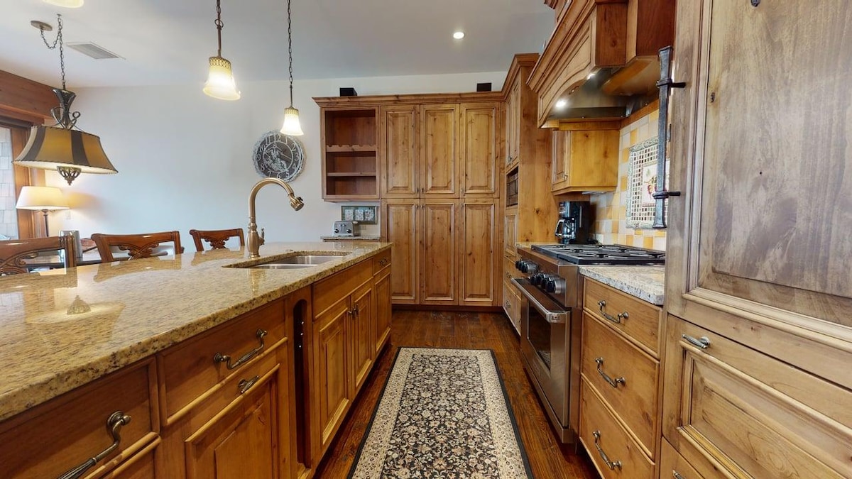 The kitchen features wooden cabinetry and granite countertops, complemented by stainless steel appliances and a gas stove. A decorative area rug is positioned on the dark wood floor, while ample cabinetry provides an organized workspace. Warm lighting fixtures hang above the island for functionality.