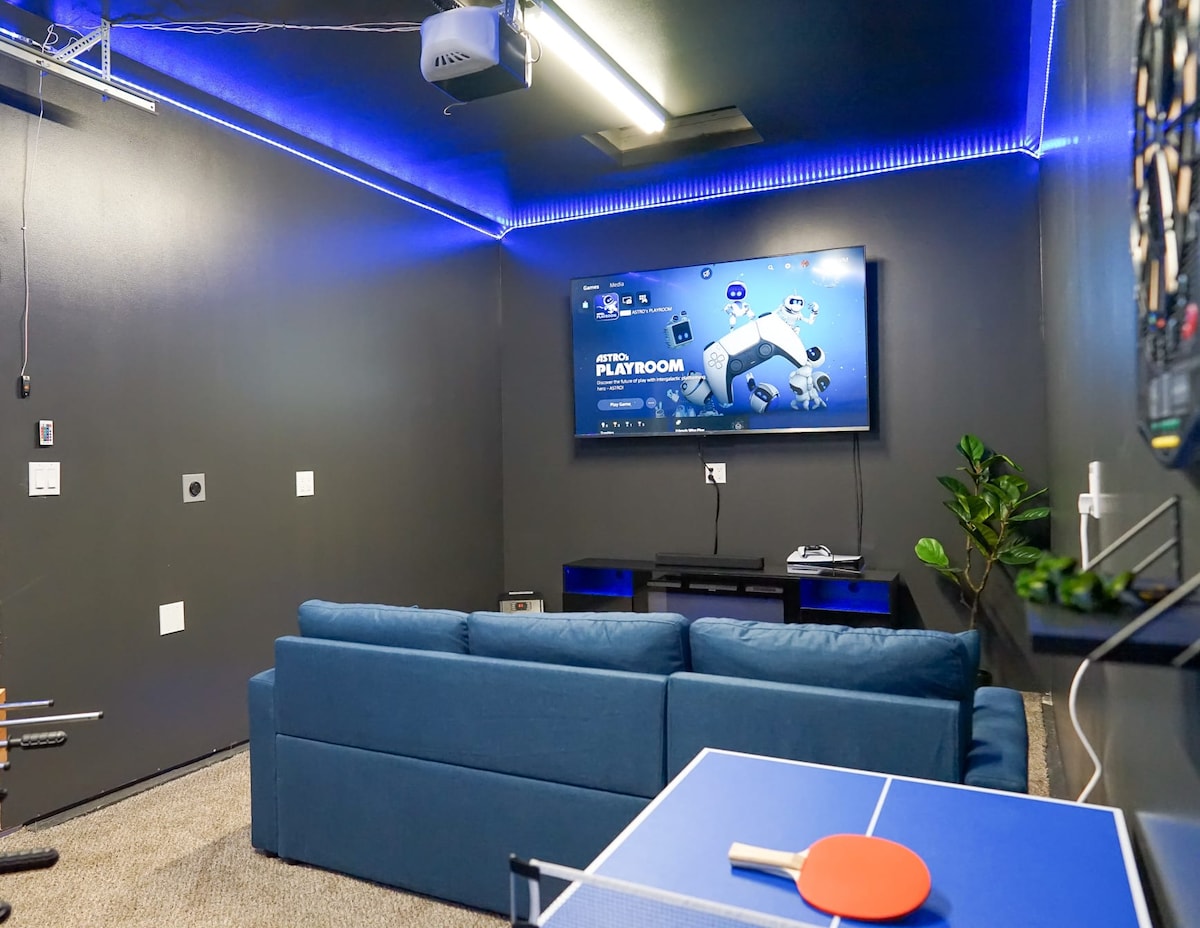 A modern entertainment room features a large Smart TV mounted on the wall, displaying a gaming interface. A comfortable blue sofa occupies the space, with a mini ping pong table and red paddle in view. LED lighting along the ceiling adds a subtle glow to the environment.