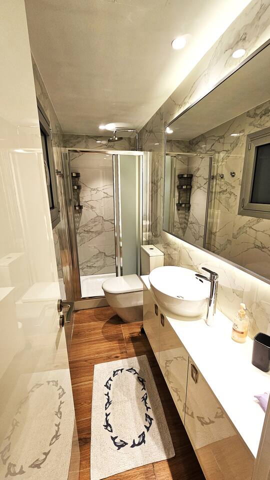 A modern bathroom features sleek marble walls and reflective surfaces. A spacious sink is situated above a long countertop, complemented by a well-lit mirror. A glass shower enclosure stands to the side, with a minimalist design enhancing the overall contemporary feel.