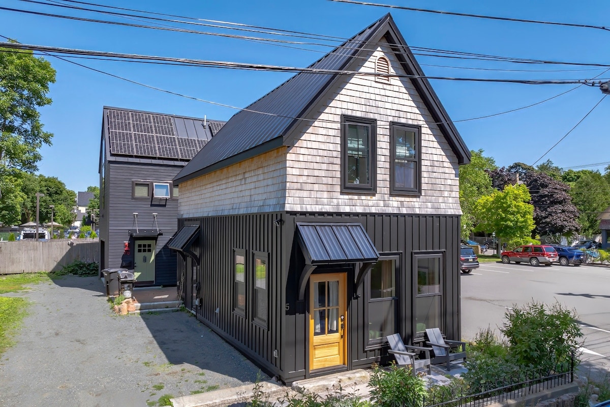 A modern, two-story building is showcased from an outdoor perspective. The exterior features a combination of black siding and wooden elements, complemented by large windows. Two chairs are positioned on the front porch, enhancing the inviting entrance, while surrounding greenery adds to the overall setting.