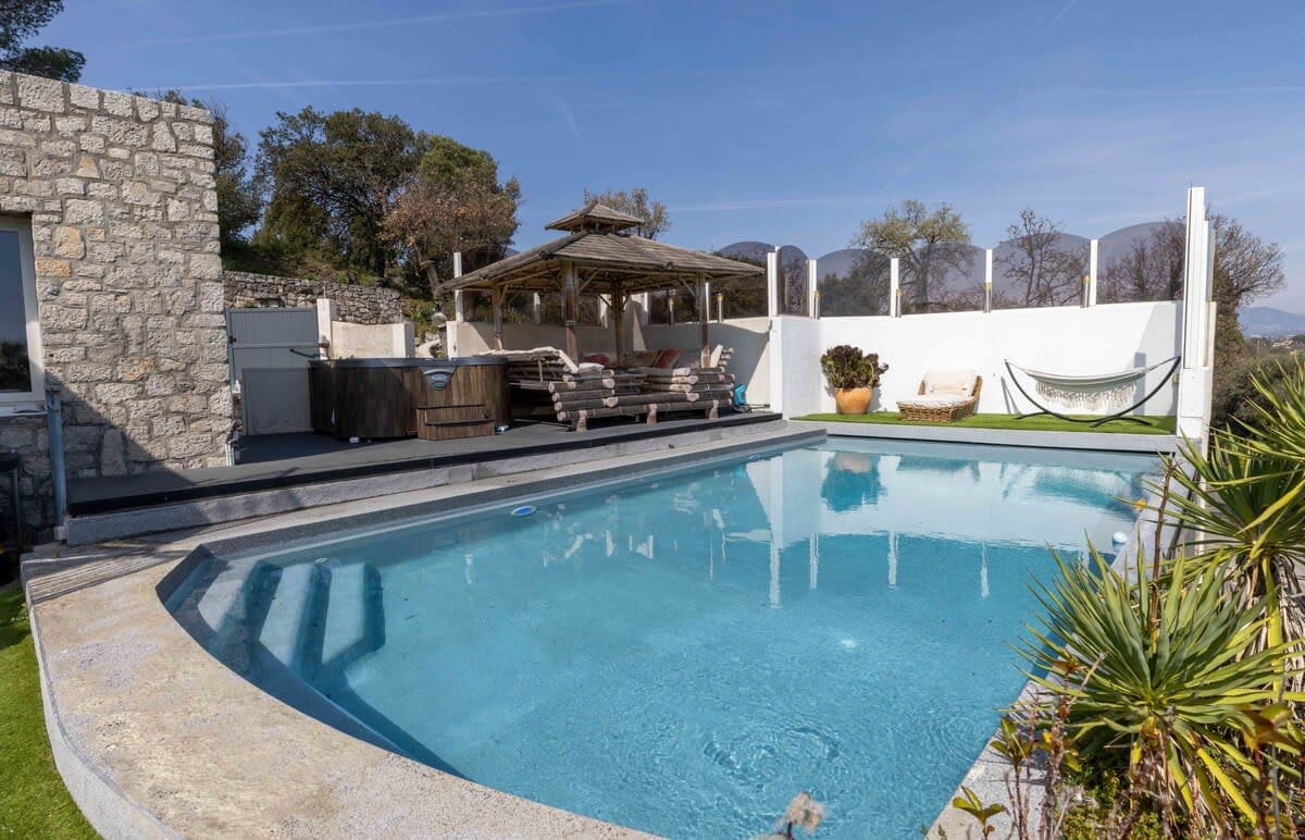 An inviting outdoor space features a clear infinity pool surrounded by smooth stone decking. Comfortable lounge chairs are situated near a shaded seating area, complemented by a hammock and potted plants. Scenic views of the surrounding landscape are visible in the background.