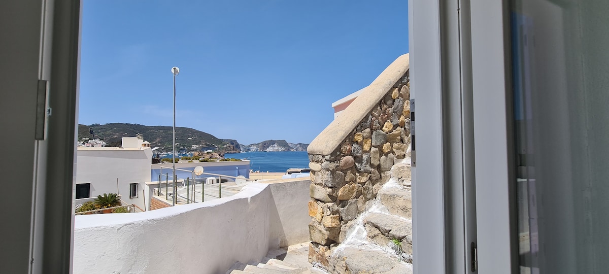 A clear view of the sea is framed by an open door, revealing a set of stone steps leading outside. The bright sky and surrounding landscape create a calm and inviting atmosphere, enhancing the connection to the scenic coastal environment.