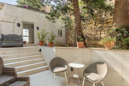 A private outdoor area features a staircase leading to a quiet entrance, bordered by lush greenery and stone walls. Two minimalist chairs and a small table are positioned on the tiled surface, providing a cozy spot for relaxation amidst the serene atmosphere.