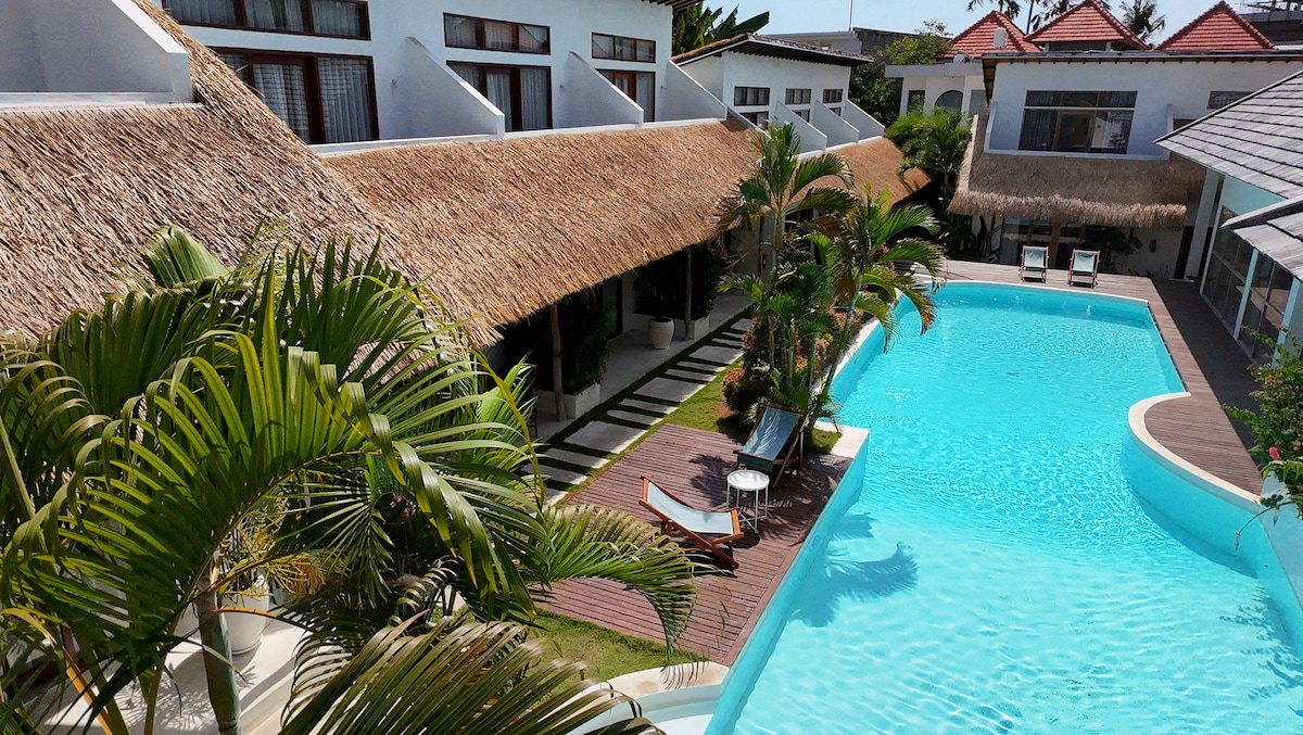 An aerial view captures a serene outdoor space featuring a large swimming pool surrounded by a wooden deck. Lush palm trees and tropical plants frame the area, while thatched-roof structures provide shade along the pathway leading to the pool, enhancing the serene atmosphere.