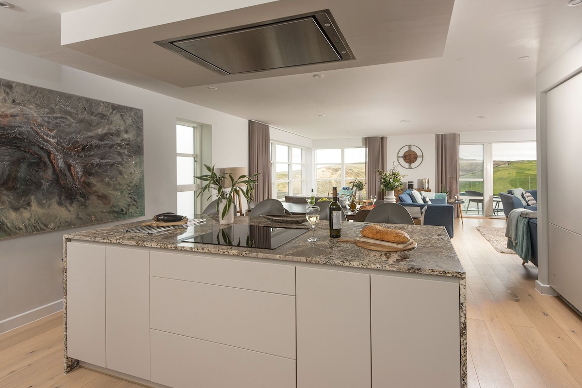 The modern kitchen features a spacious island with a granite countertop, equipped with a built-in induction hob. A large painting adorns the wall, while the open layout connects to the living area, showcasing large windows that offer views of the surrounding scenery.