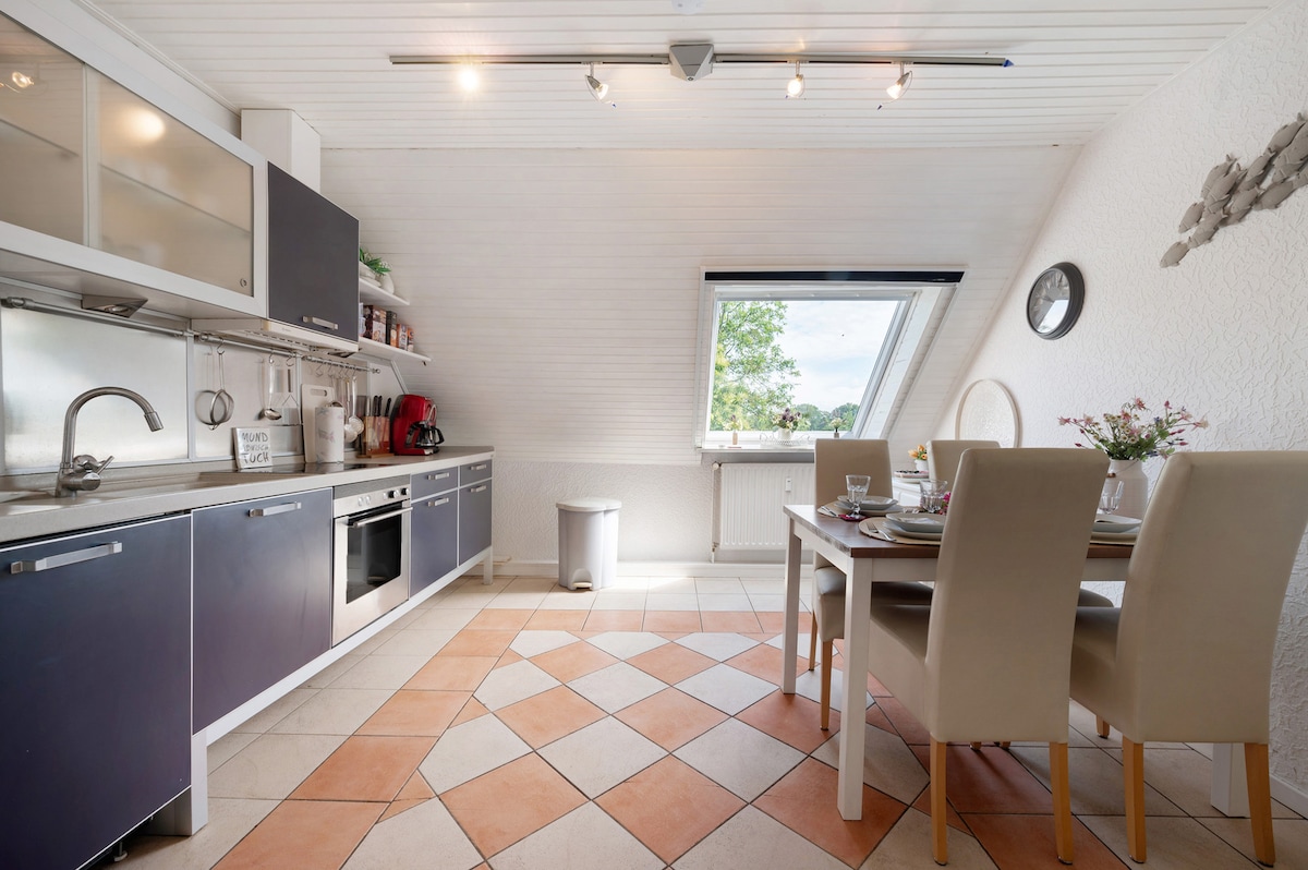 A well-equipped kitchen is displayed, featuring dark cabinetry and a silver oven. Natural light fills the space through a large window, which overlooks greenery. A dining table with four upholstered chairs is set for a meal, complemented by a decorative centerpiece.