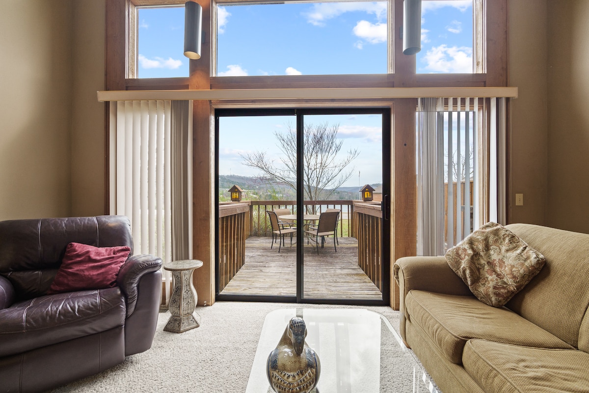A spacious living area features plush seating arrangements, including a dark leather couch and a light-colored sofa. Natural light floods in through large glass doors that lead to a private deck with outdoor seating, offering views of the serene lake and surrounding landscape.