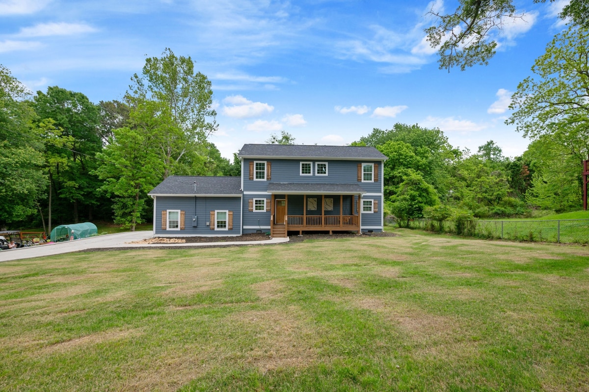 A two-story home features a gray exterior and a welcoming front porch. Surrounded by lush greenery, the property offers a spacious lawn and ample outdoor space. A paved driveway leads up to the entrance, with trees lining the background for added privacy.
