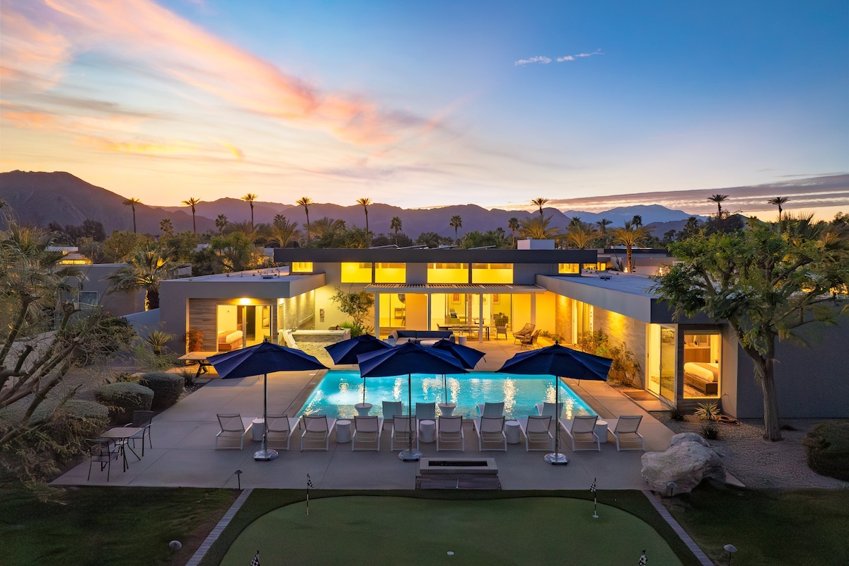 An aerial view captures a modern home with a spacious pool surrounded by lounge chairs and large umbrellas. The exterior is highlighted by warm lighting from within, and palm trees frame the expansive outdoor space, set against a stunning sunset backdrop.