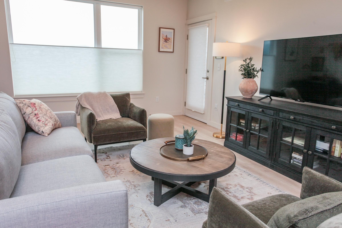 A modern living area displays a comfortable sectional sofa and an accent chair. A round coffee table with a plant centerpiece sits in front of a television mounted above a black media console. Large windows with light-filtering shades provide natural light.