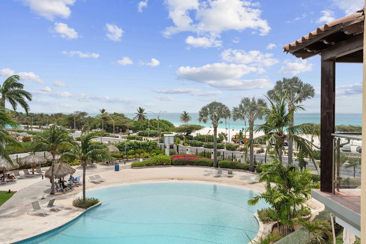 A panoramic view of the resort's circular swimming pool is showcased, surrounded by lush landscaping and lounge chairs. Beyond the pool, the pristine white sand of Eagle Beach is visible under a clear sky, framed by palm trees that sway gently in the breeze.