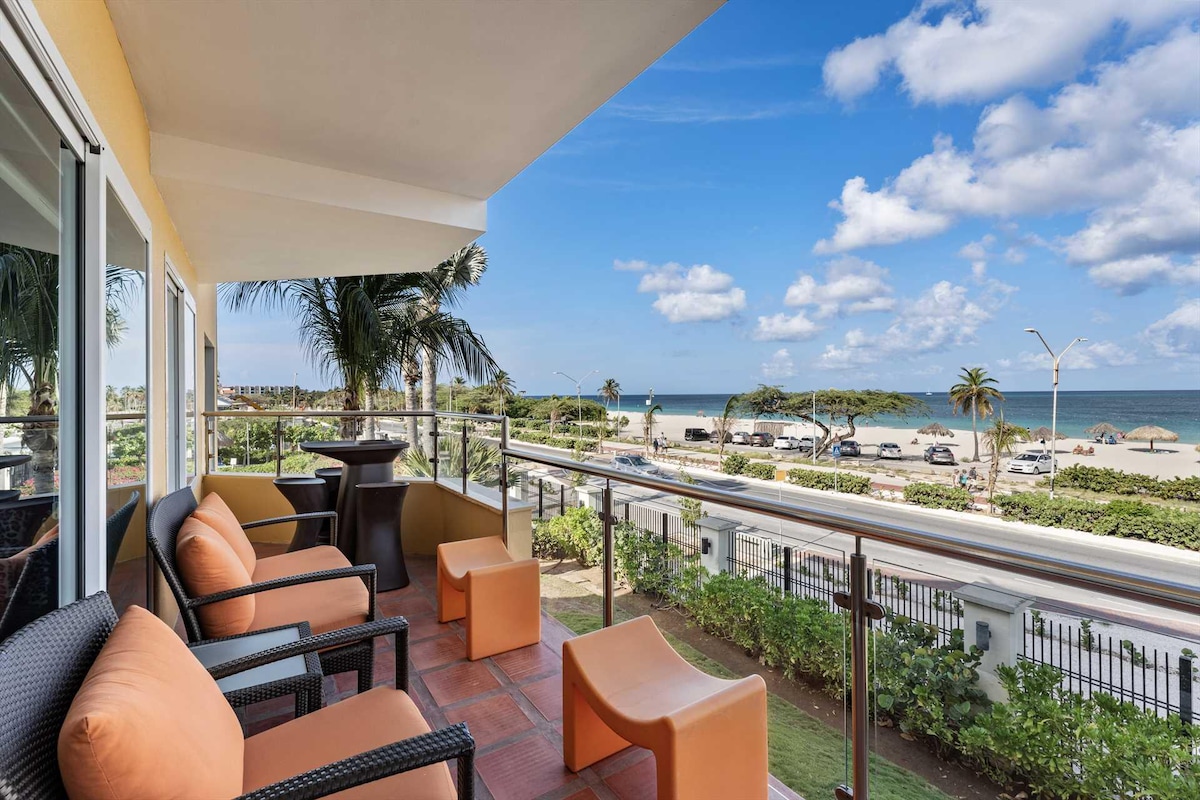 An inviting balcony is furnished with lounge chairs and small tables, offering unobstructed views of the beach and ocean. Lush green landscaping and palm trees frame the scene, while a clear blue sky and soft clouds provide a serene backdrop.