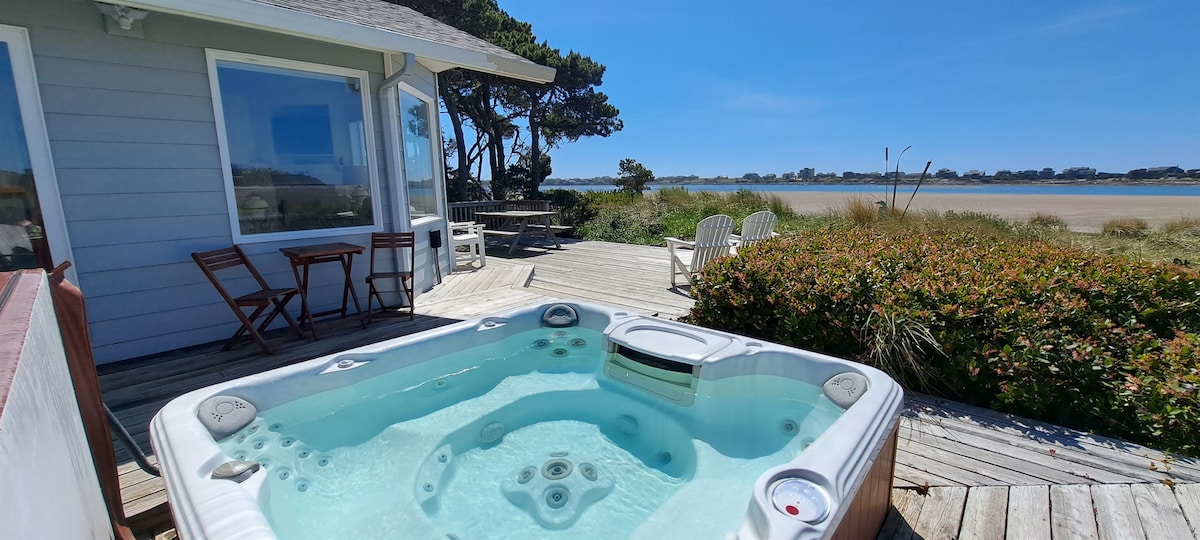 A hot tub is positioned on a wooden deck, offering views of the tranquil waterfront. A small table with two chairs is visible nearby, alongside additional seating that enhances the outdoor space. Lush greenery frames the area, complementing the clear blue sky.