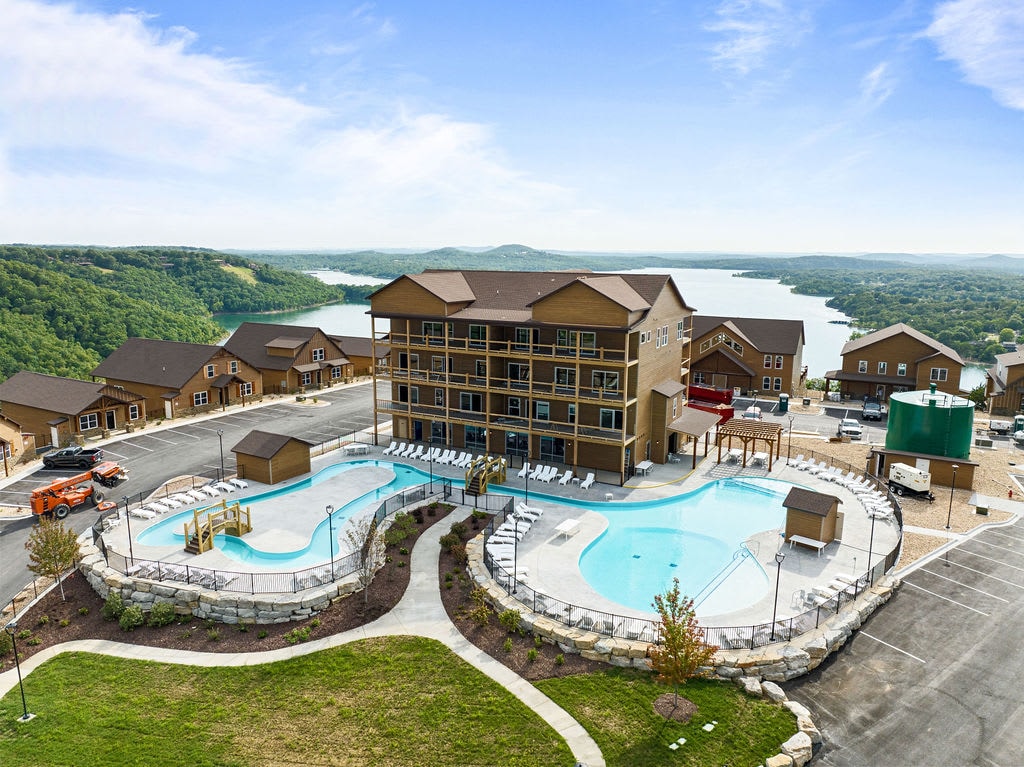 The image captures a community pool area surrounded by a spacious outdoor deck. The pool features a lazy river design, complemented by lounge chairs positioned around it. In the background, additional accommodations alongside scenic views of Table Rock Lake and green hills are visible.