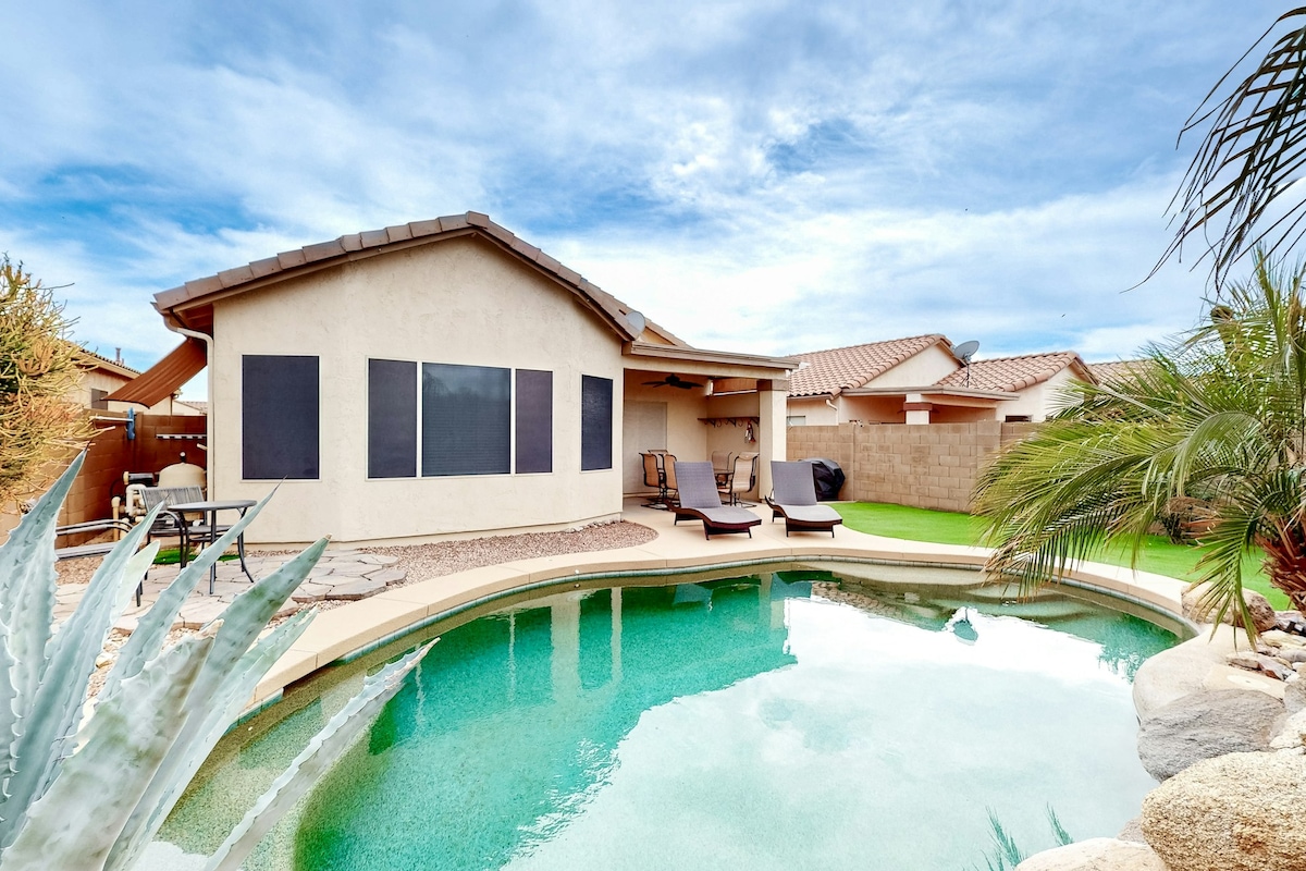 The image captures a serene backyard with a private pool, reflecting the sky's hues. A single-story house features large windows on one side and a covered patio with seating. Lush green grass surrounds the pool, complemented by desert landscaping and palm trees.