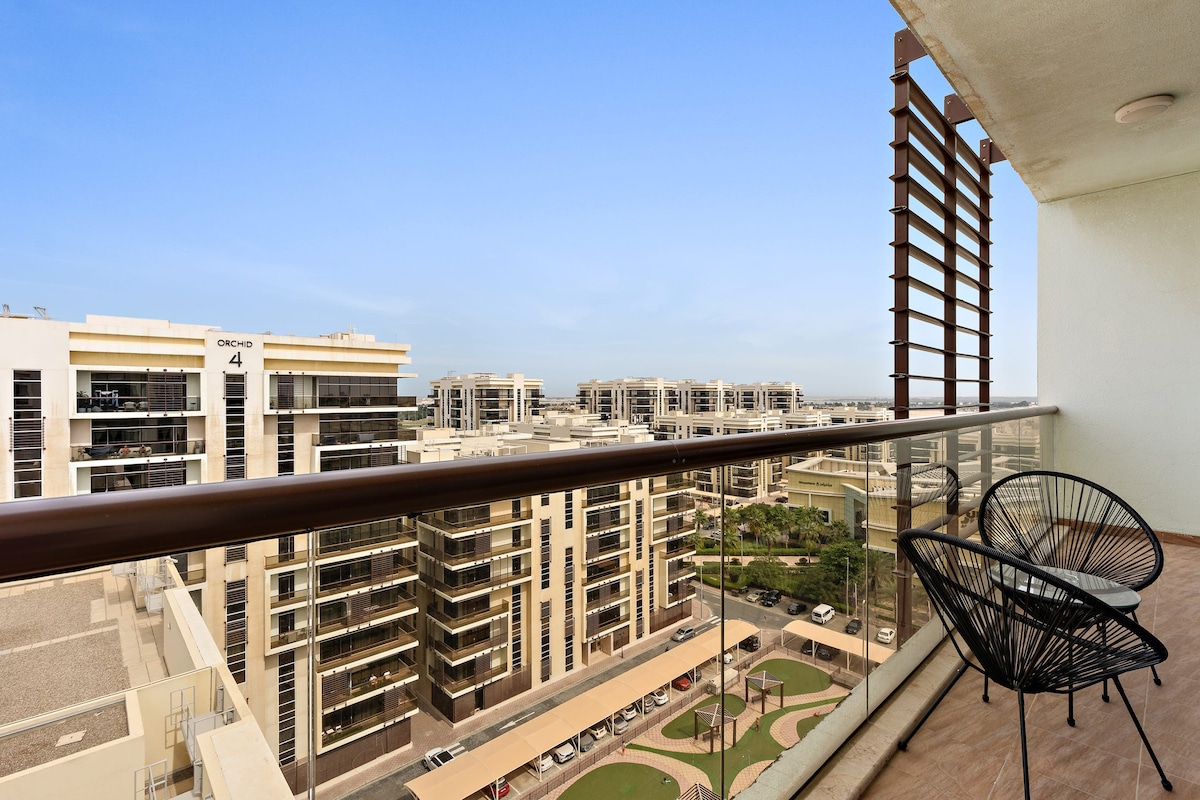 A spacious balcony is depicted, featuring two stylish chairs overlooking the community landscape. Below, green areas and pathways are visible among modern apartment buildings, creating an open and airy atmosphere with a clear blue sky providing a bright backdrop.