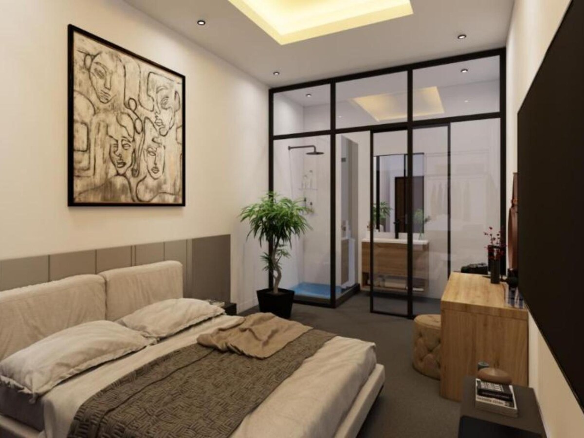 A modern room features a spacious bed with neutral linens and a decorative blanket. A sleek wooden dresser and a potted plant complement the space. Glass partitions reveal an ensuite bathroom. Soft lighting enhances the overall ambiance, while an artwork adorns the wall.