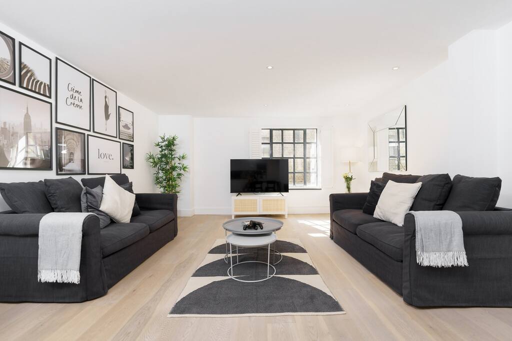The open-plan living room features two dark gray sofas positioned across from each other, complemented by a round coffee table in the center. A large 58-inch TV is mounted on the wall, with decorative black and white framed images nearby. Natural light fills the space through large windows.