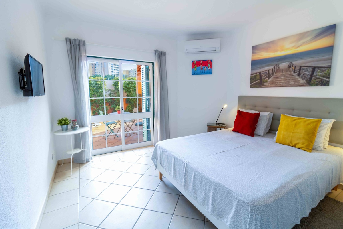 A well-lit room features a spacious bed adorned with colorful cushions. A wall-mounted TV is positioned opposite the bed. Large glass doors open to a balcony, allowing ample natural light. A small table and chairs are set up outside, enhancing the outdoor experience.