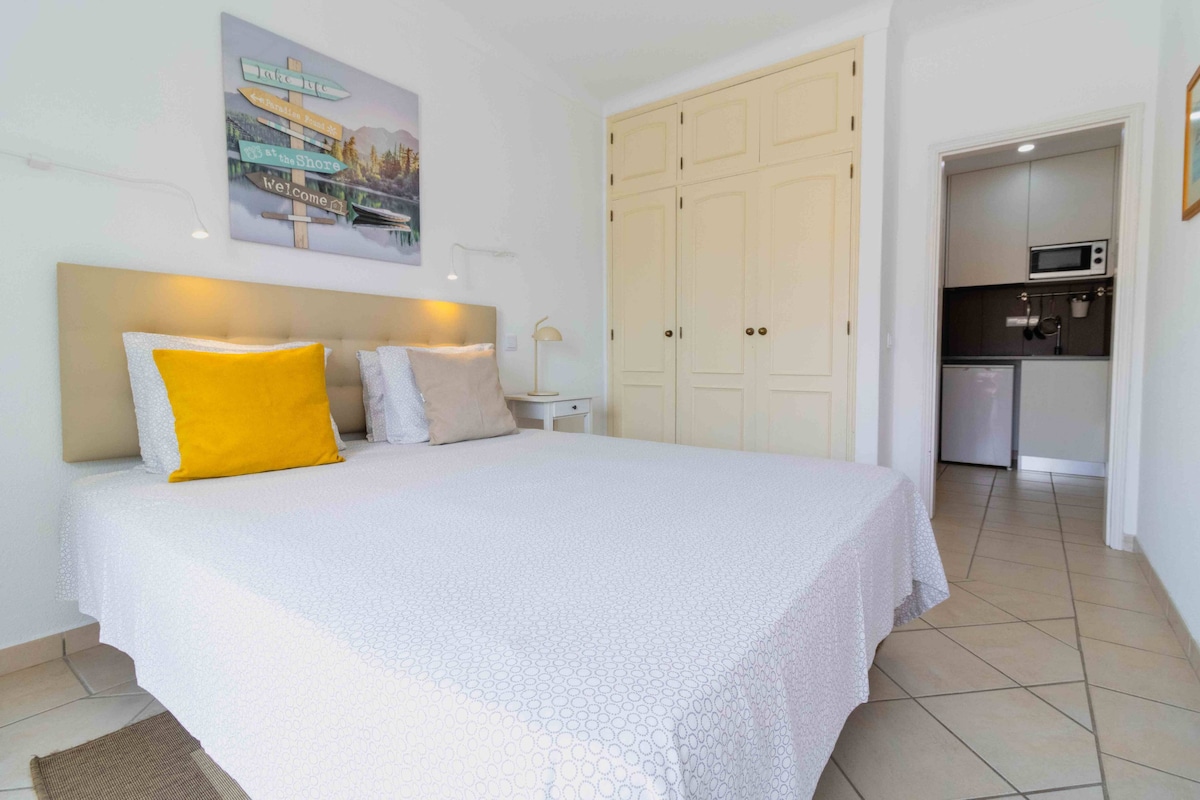 A spacious bedroom features a comfortable double bed with decorative pillows in muted tones. Built-in cabinets provide ample storage. A small kitchenette is visible in the background, equipped with a microwave and a mini fridge, enhancing the room's practicality.