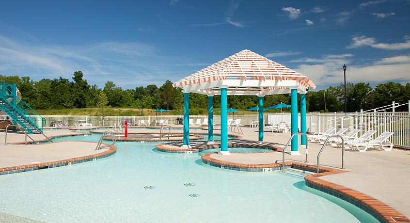 A refreshing outdoor pool area is featured, with clear blue water and a gradual slope for easy access. A shaded cabana provides respite from the sun, and lounge chairs are arranged around the deck for relaxation. A water slide is visible in the background.