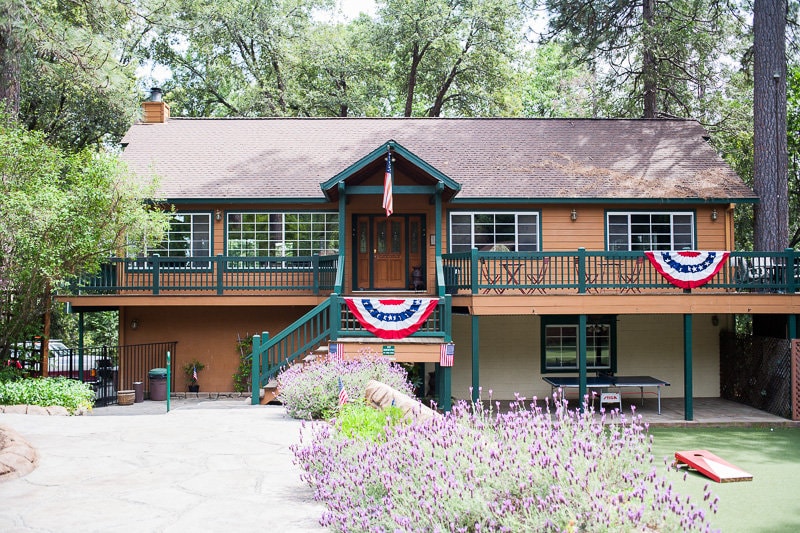 The exterior of the two-story home is surrounded by lush greenery, showcasing a welcoming entrance with steps leading to a spacious porch. Red, white, and blue decorations adorn the railings, and vibrant flowers add color to the inviting landscape.