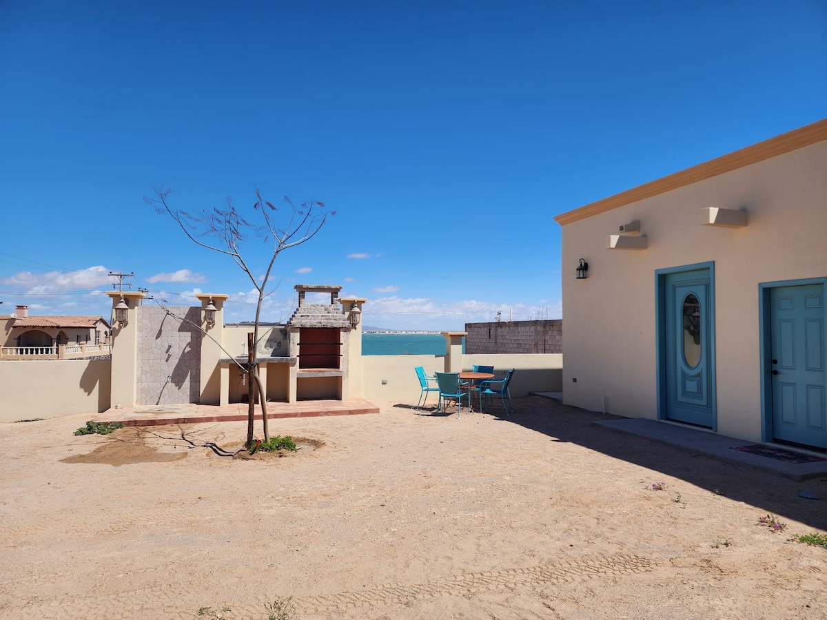 An open outdoor space features a sandy area with minimal vegetation and a small tree. A covered barbecue grill is situated against a wall, alongside a table with four blue chairs, offering views of the distant sea under a clear blue sky.