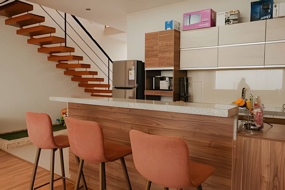 A modern kitchen area is highlighted by a sleek countertop and wooden cabinetry, featuring a refrigerator and kitchen appliances. Three bar stools with soft upholstery are positioned at the counter. A staircase with wooden steps leads to a mezzanine level, enhancing the open layout.