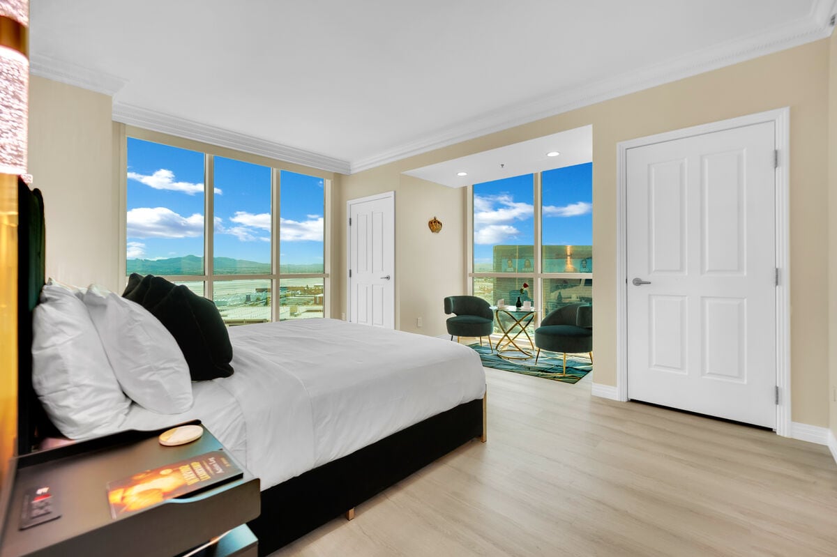 A bright sleeping area showcases a plush king bed with crisp white linens. Large windows allow for natural light, revealing views of the distant mountains and cityscape. A cozy seating area is positioned beside the windows, creating a serene space for relaxation.
