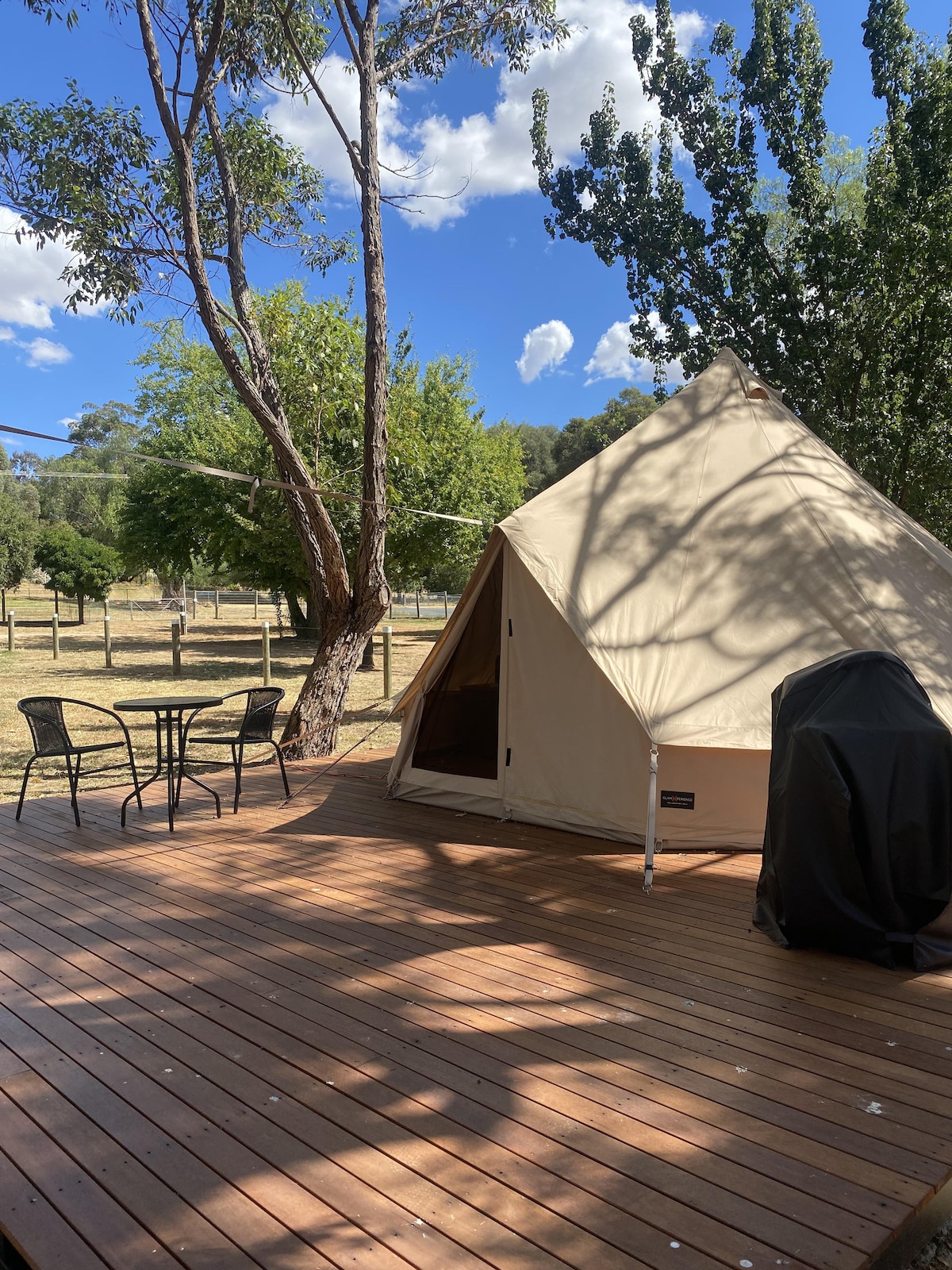 A glamping tent is set on a wooden deck, surrounded by greenery. A small table with two chairs is positioned nearby. Sunlight casts shadows from the trees, creating a natural ambiance. The tent's entrance is visible, leading into the sleeping area.