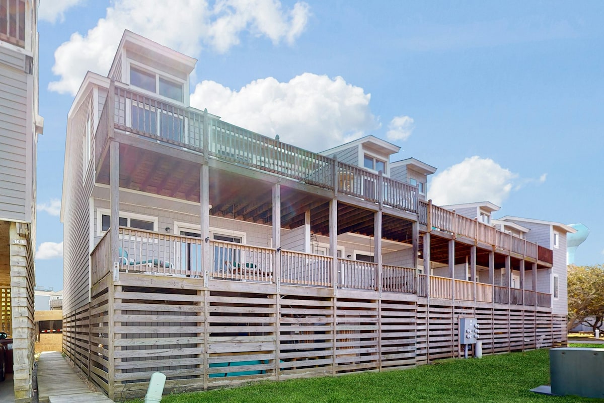 The three-story townhouse features expansive decks on each level, allowing for outdoor relaxation. The wooden railings and open design offer a spacious feel, while green lawn provides a contrasting natural setting. The building showcases multiple windows, ensuring ample sunlight throughout the home.
