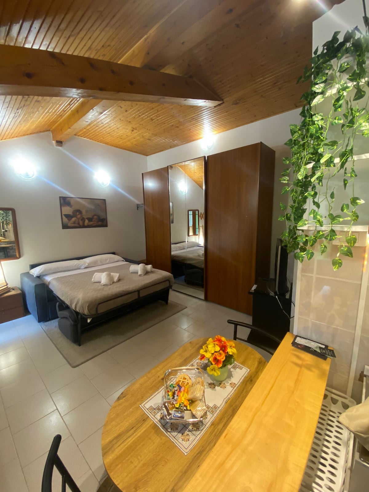 A spacious monolocale is showcased with wooden beams overhead and natural light from the windows. A comfortable bed is arranged with fresh linens and decorative towels. A dining table is positioned nearby, adorned with a floral centerpiece, while a wardrobe is reflected in the mirror.