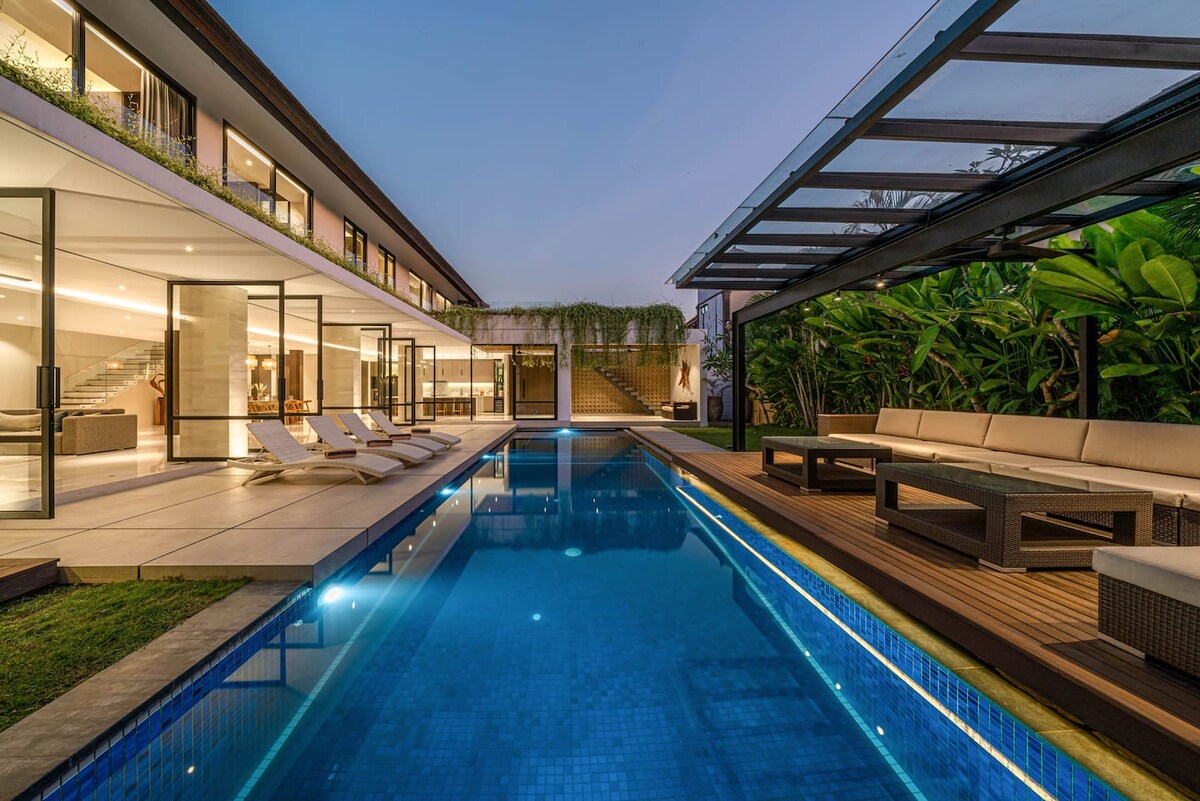An expansive pool is framed by lush tropical gardens, with comfortable lounge chairs positioned nearby. Floor-to-ceiling glass doors provide a view of the modern villa's interior, featuring a light-filled open layout. Subtle outdoor lighting enhances the serene ambiance as evening approaches.