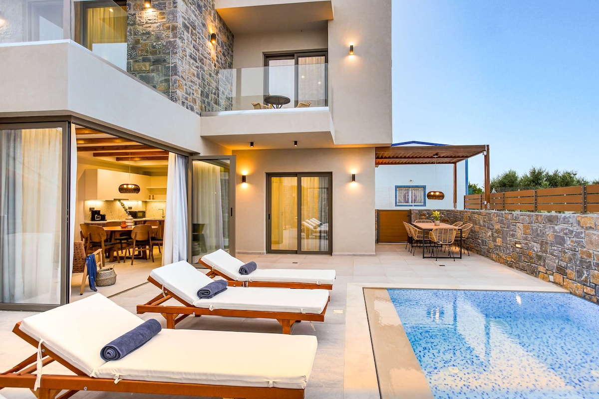A private pool area features two sun loungers with rolled towels, set against a backdrop of a modern villa. The glass doors open to an inviting living space, while a dining area is visible in the distance. Soft evening light casts a serene ambiance.