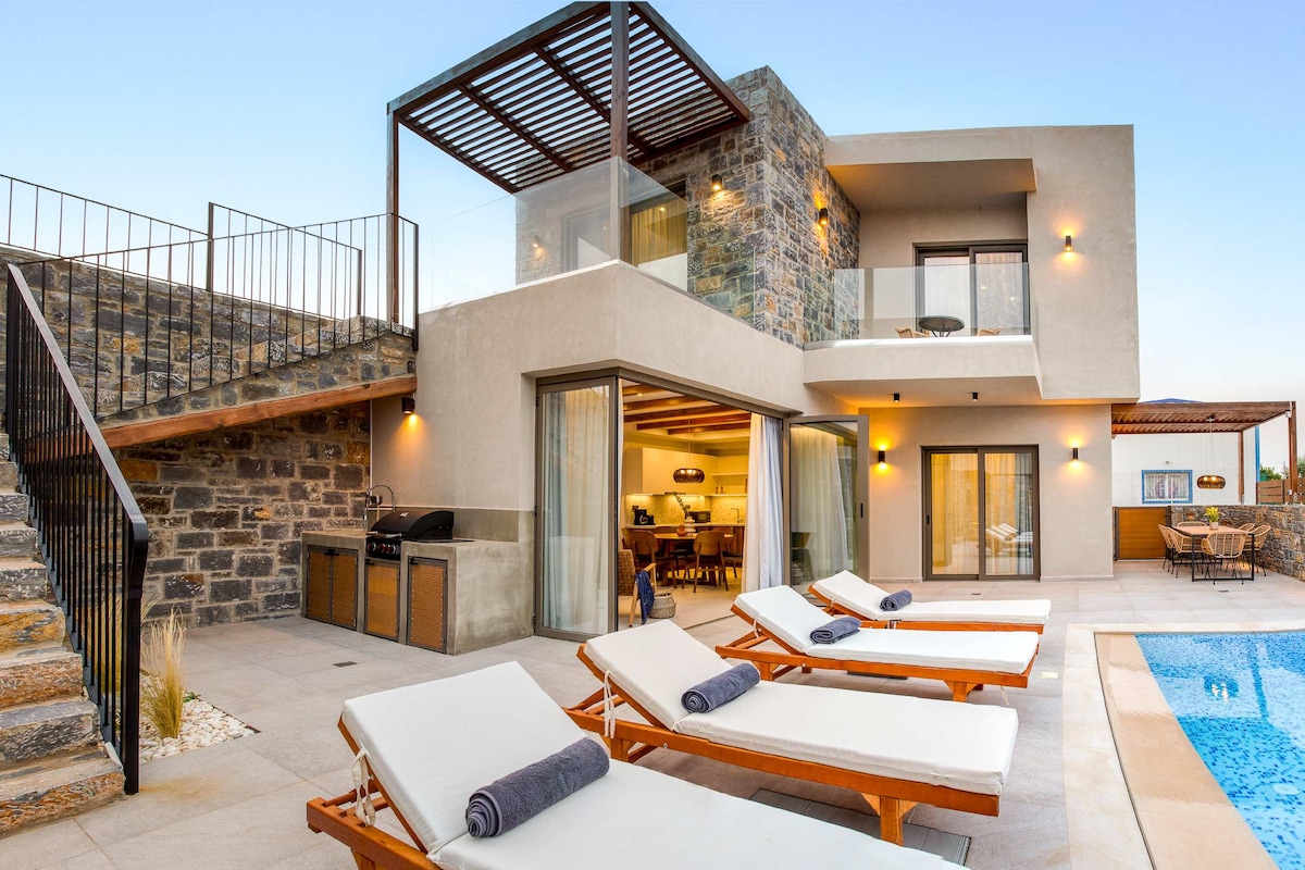 The two-story villa features a modern exterior with stone accents and large windows. Sun loungers are arranged beside the private pool, while a BBQ grill is visible on the patio. Stairs lead to an upper-level balcony, enhancing the outdoor living space.