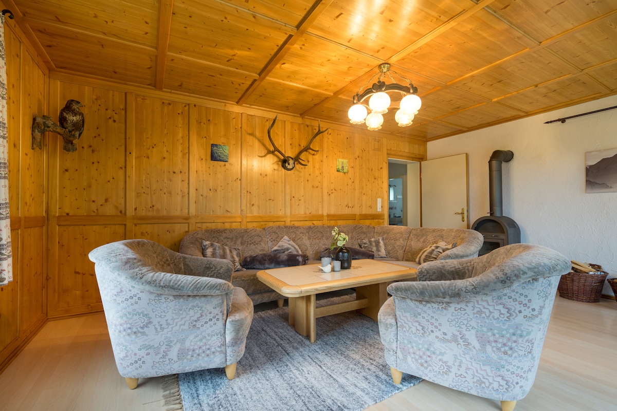 A spacious living area features wooden paneling and a cozy seating arrangement with soft armchairs. A wooden coffee table is centered on a textured rug, while ambient light from a unique chandelier illuminates the room. A small wood stove is positioned in the corner.
