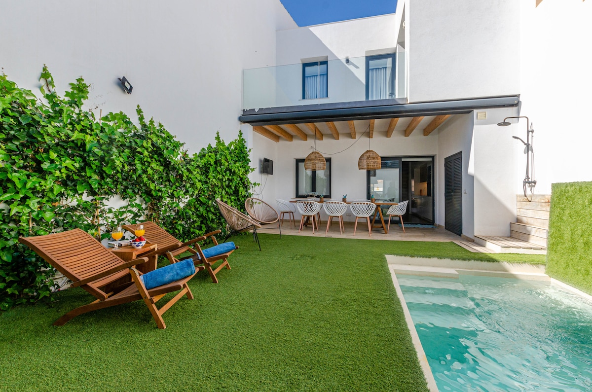 The outdoor area showcases a private saltwater pool adjacent to a garden with artificial grass. Two sun loungers are positioned for relaxation, while a dining table is visible in the background. Lush greenery adds a touch of nature to the tranquil setting.