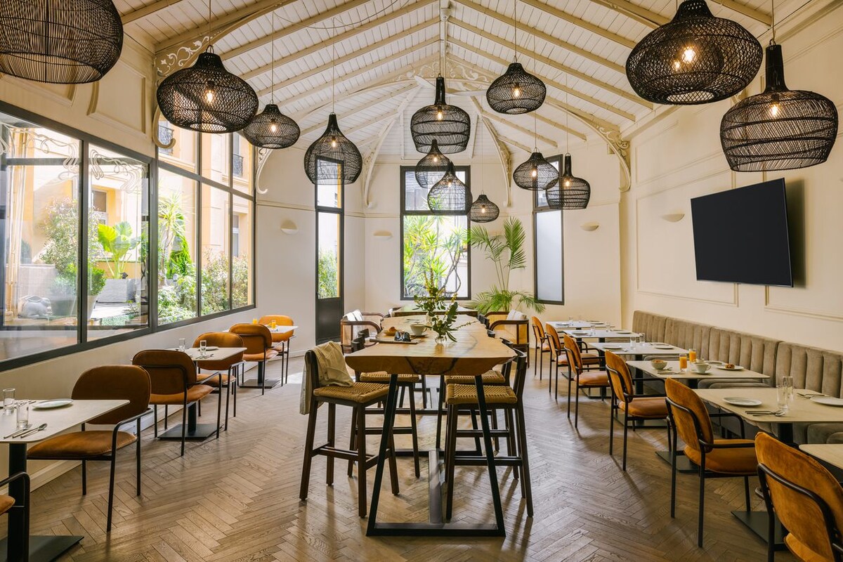 An inviting dining space features a mix of upholstered chairs and high stools surrounding a large communal table. Large windows allow natural light to fill the room, enhancing the greenery outside. Unique pendant lights hang from the ceiling, adding character to the minimalist decor.