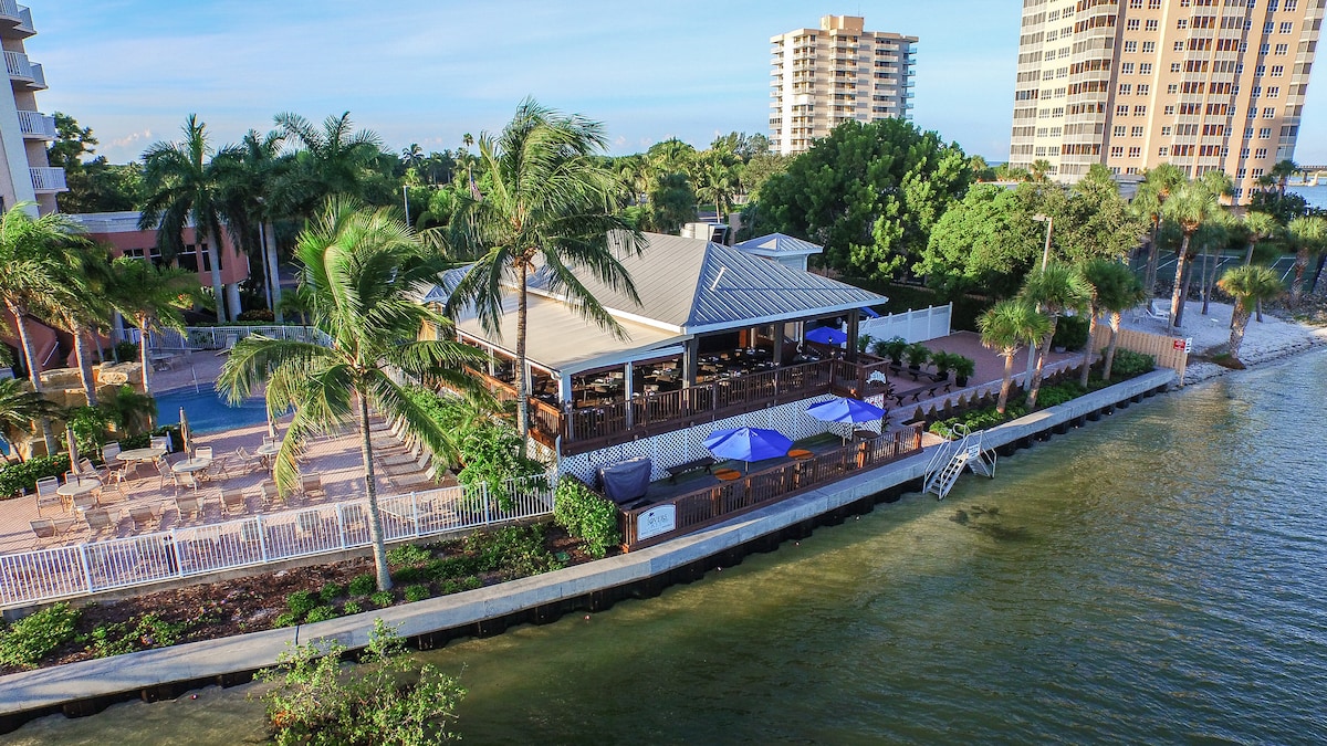 A waterfront view is presented, showcasing a restaurant structure with a large open deck surrounded by palm trees. Umbrellas provide shade for seated areas, while a pathway leads to the water's edge. Nearby buildings offer a backdrop of residential life.
