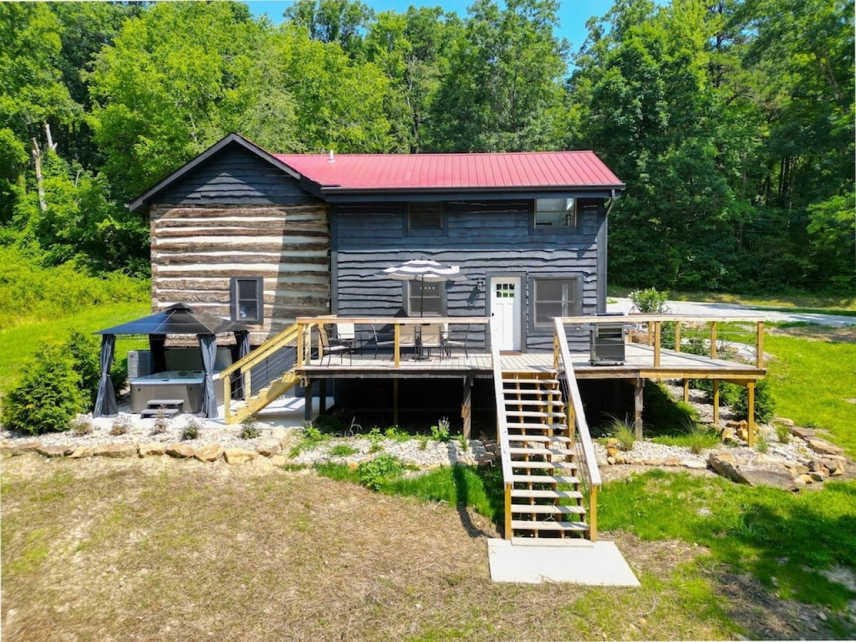 The rustic cabin features a wooden deck with steps leading up to the entrance. A shaded seating area is positioned beside the cabin, with parasols providing cover. Lush greenery surrounds the home, enhancing its secluded setting in nature.