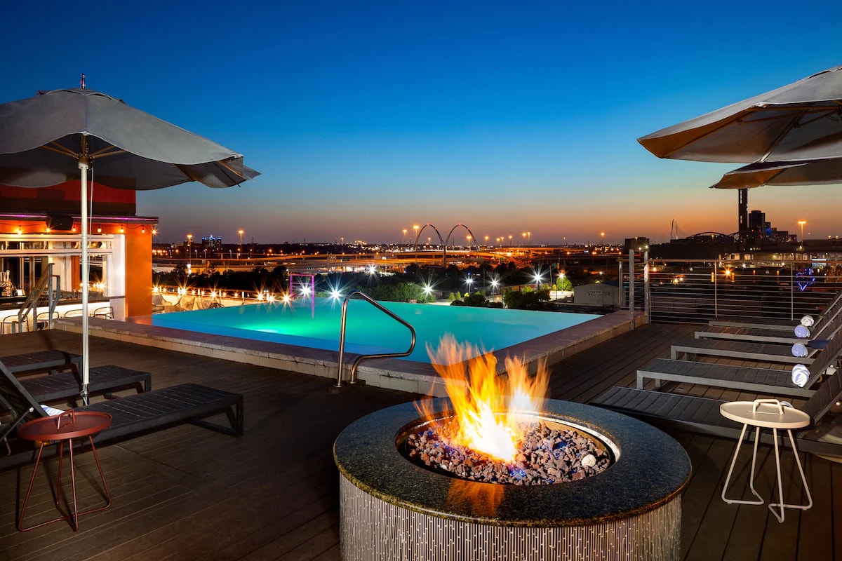 A rooftop oasis is highlighted by a sparkling pool that reflects the twilight sky. Comfortable lounge chairs surround a modern fire pit, where flames dance over decorative stones. Umbrellas offer shade while the city skyline provides a captivating backdrop.