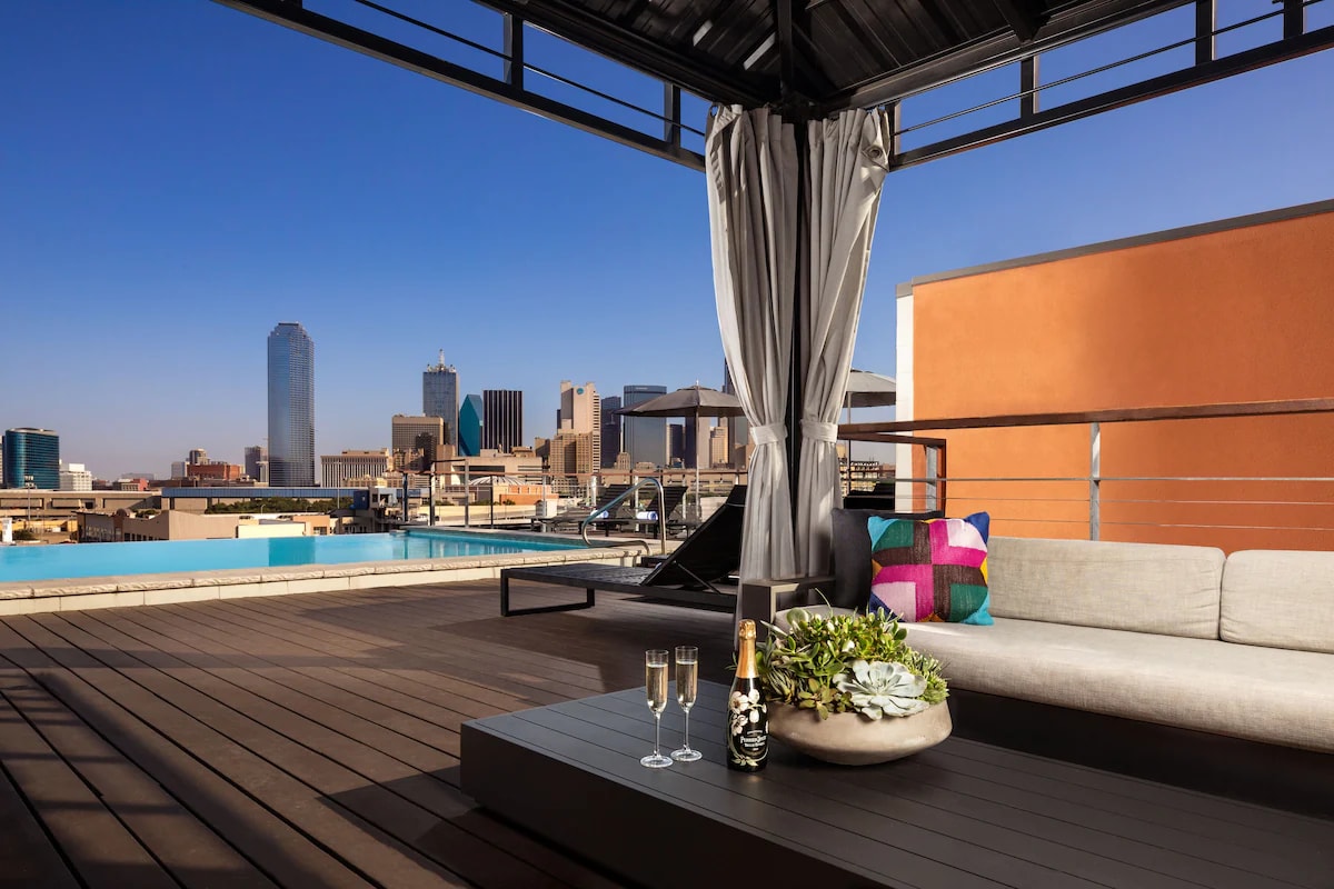 A rooftop lounge area features a spacious sofa and a low table, overlooking the Dallas skyline. Soft curtains billow in the breeze, while an arrangement of greenery and two glasses of champagne sit on the table, suggesting an inviting space for relaxation.