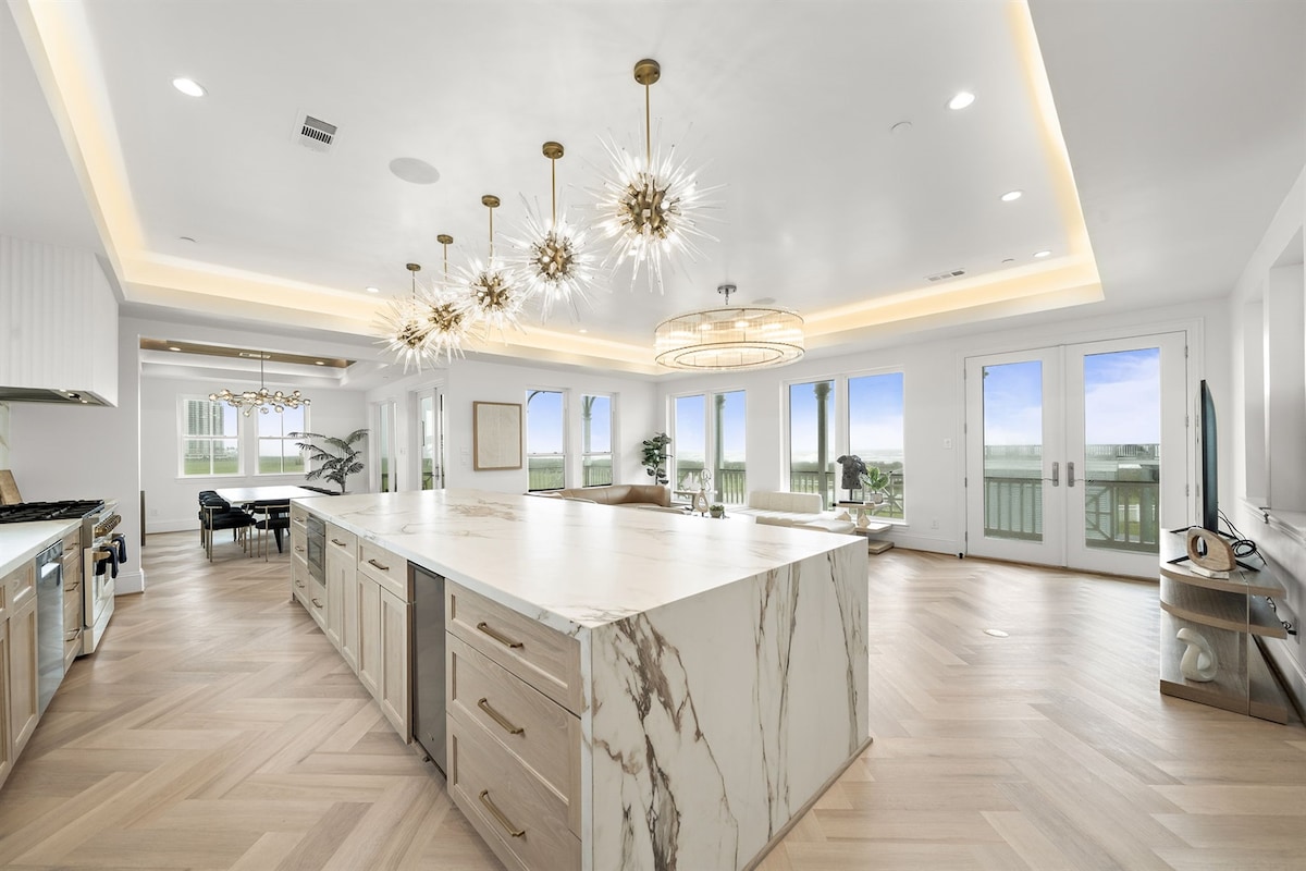 An open-concept kitchen and dining area is illuminated by modern light fixtures. A large marble island with ample counter space is central to the room. Floor-to-ceiling windows offer expansive views of the Gulf, while elegant glass doors provide access to outdoor seating.