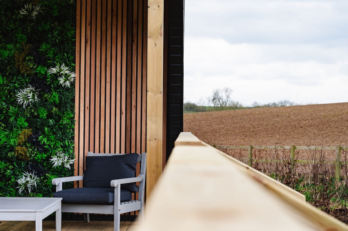 A private decking area is seen, featuring a sturdy wooden railing and a comfortable chair positioned near a small table. Behind the seating, a lush green wall adds a touch of nature, while expansive countryside views are visible in the background.