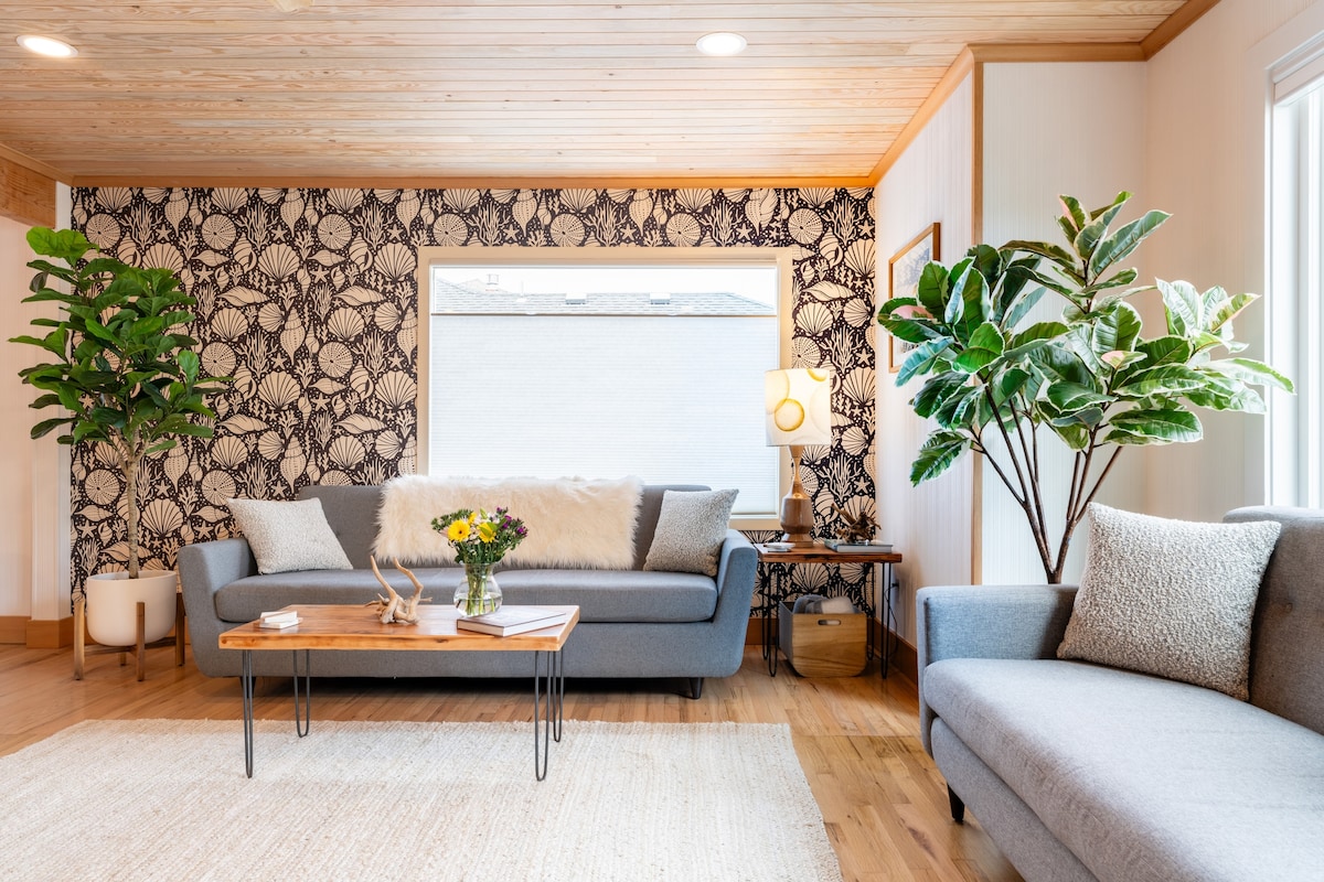 A cozy living area is featured, showcasing two gray sofas with neutral throw pillows. A wooden coffee table sits in the center, accompanied by decorative pieces. Bright greenery from potted plants enhances the space, while a patterned wallpaper adds a touch of coastal charm.