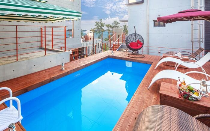 A private pool is surrounded by a wooden deck, featuring two lounge chairs and a circular hanging chair. The pool's vibrant blue water reflects the sky, while an umbrella and striped awning provide shaded seating areas nearby.