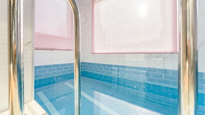 A private pool area features sleek blue tiles and is enclosed by transparent glass. Natural light filters through partially closed blinds, illuminating the water's surface and creating a serene environment.